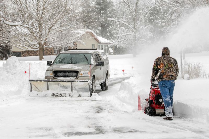 Worker in winter gear clearing a residential driveway with a snow blower while a plow truck waits in the background